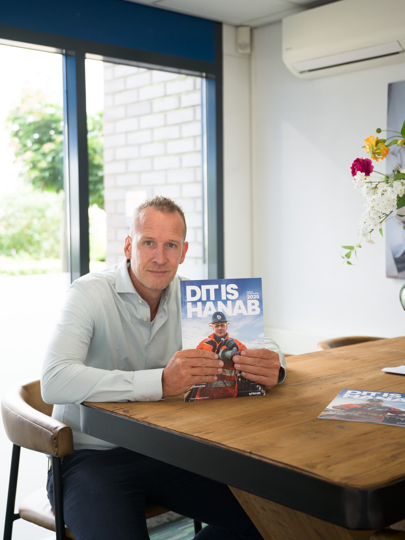 Man houdt Hanab jubileummagazine 'Dit is Hanab 2025' vast aan houten tafel op kantoor