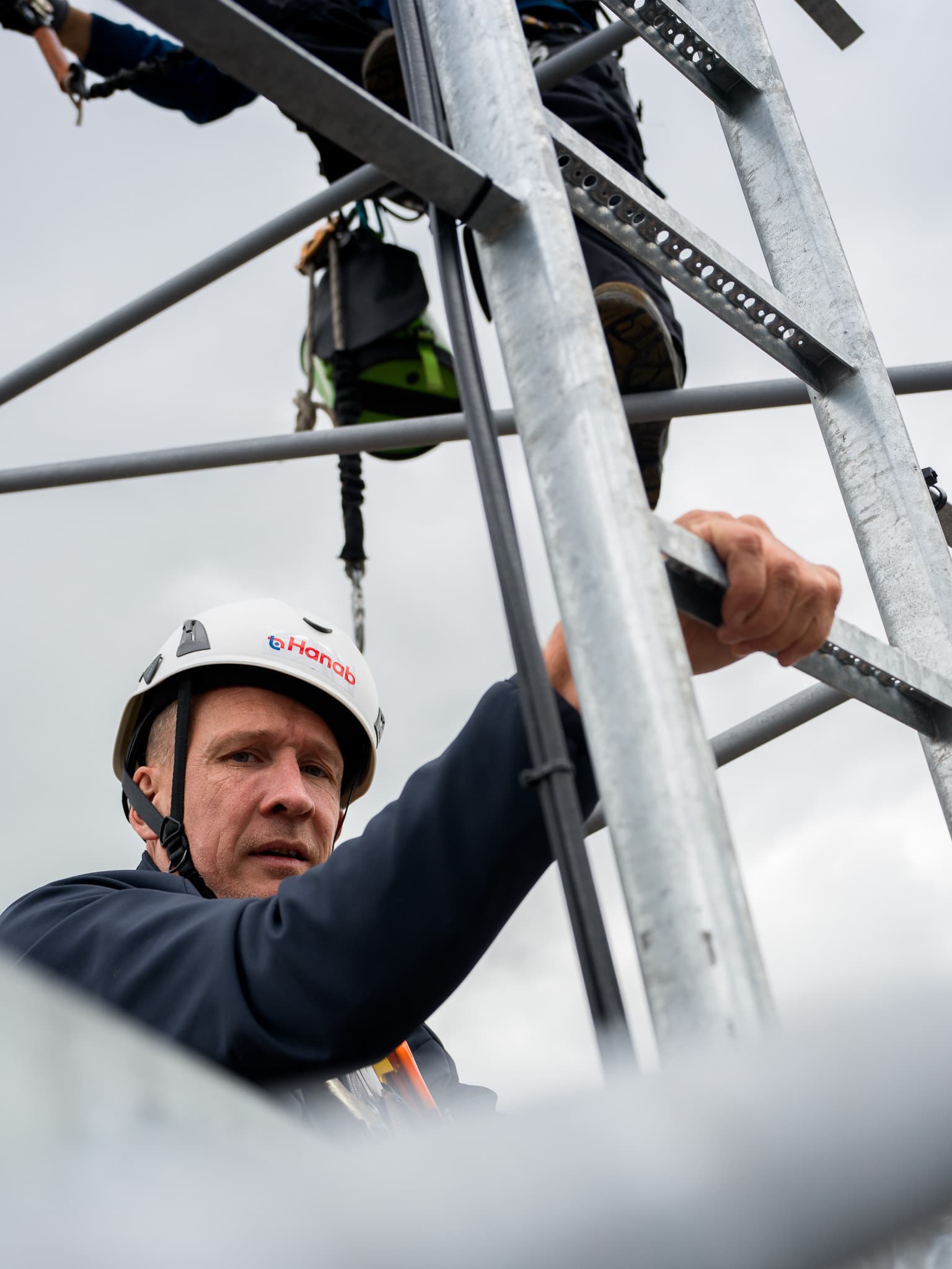 Monteur met Hanab-helm klimt in zendmast tussen stalen tralieconstructie tegen bewolkte lucht