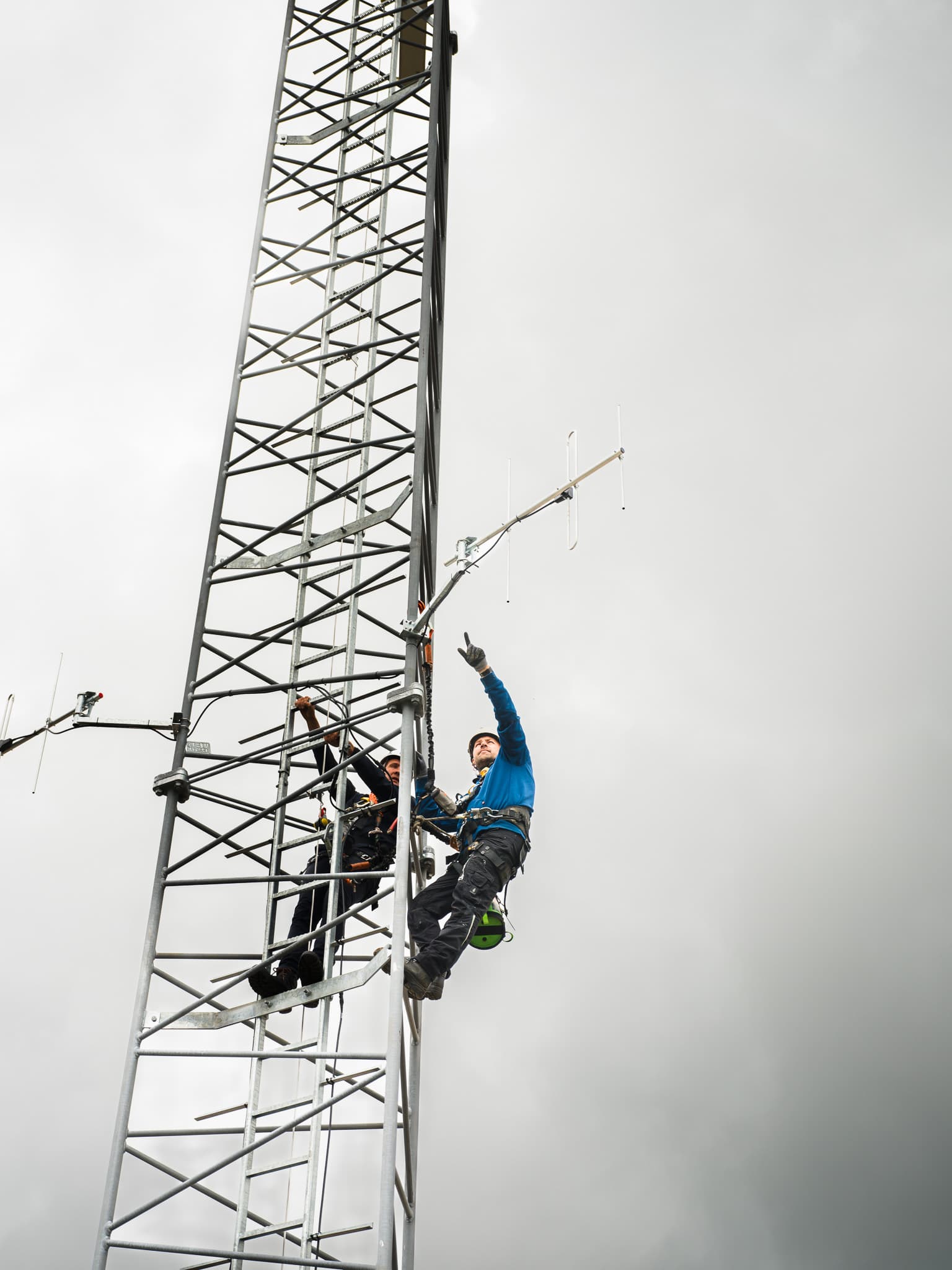 Twee monteurs beklimmen samen een hoge zendmast met antennes tegen grijze lucht
