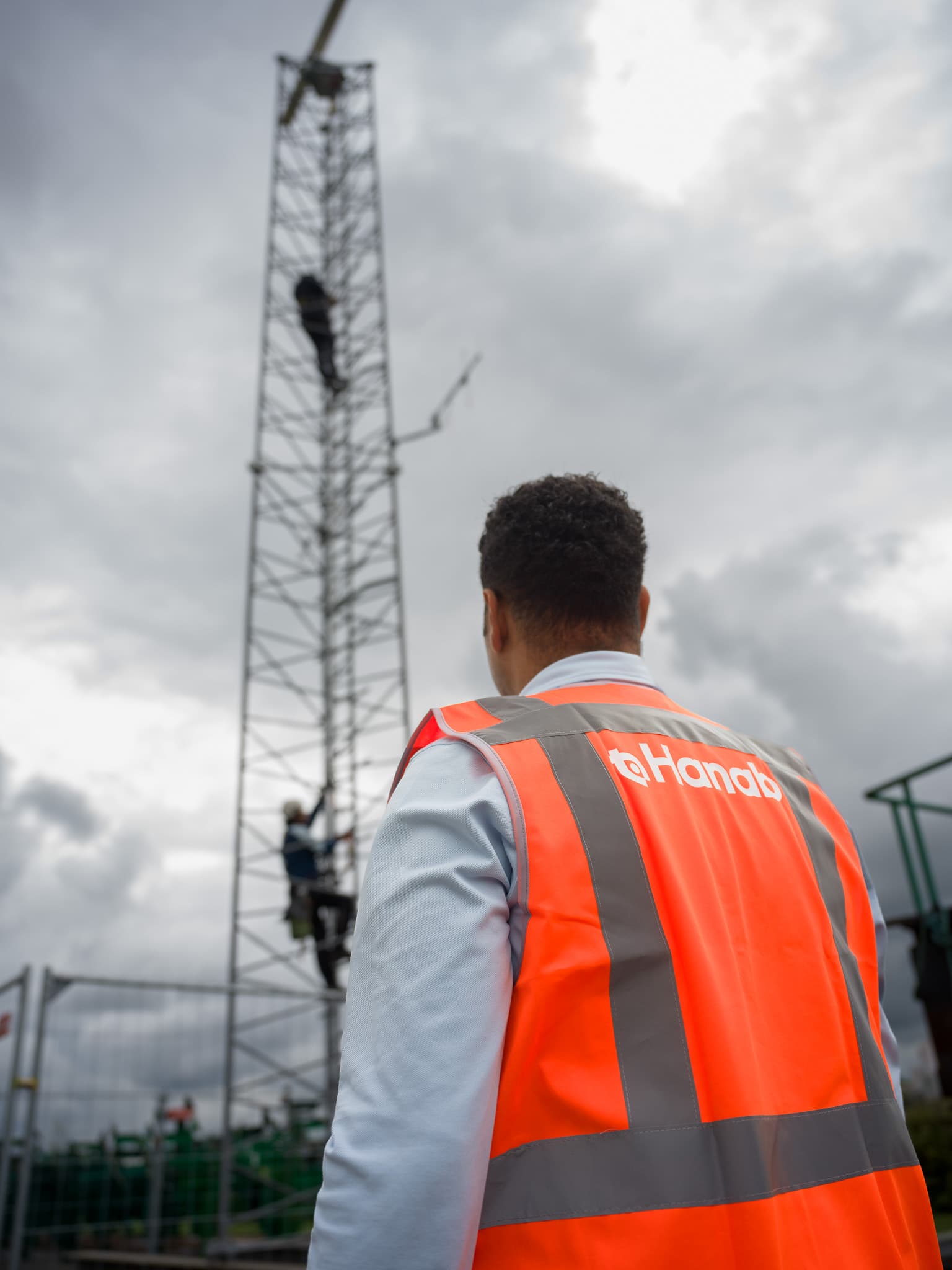 Medewerker in Hanab-vest kijkt omhoog naar collega's die zendmast beklimmen op locatie