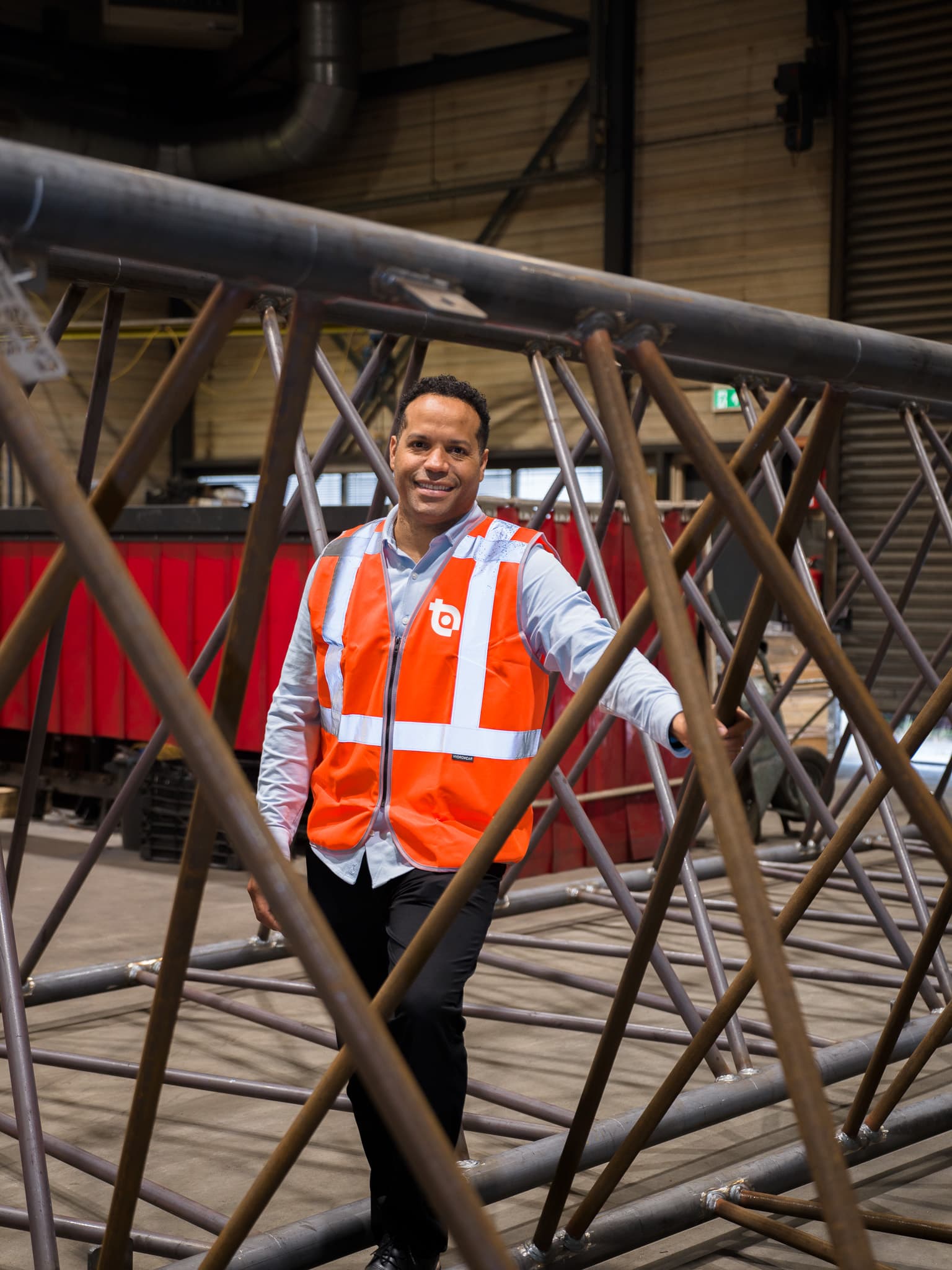 Medewerker in oranje veiligheidsvest poseert tussen grote stalen mastconstructie in fabriekshal