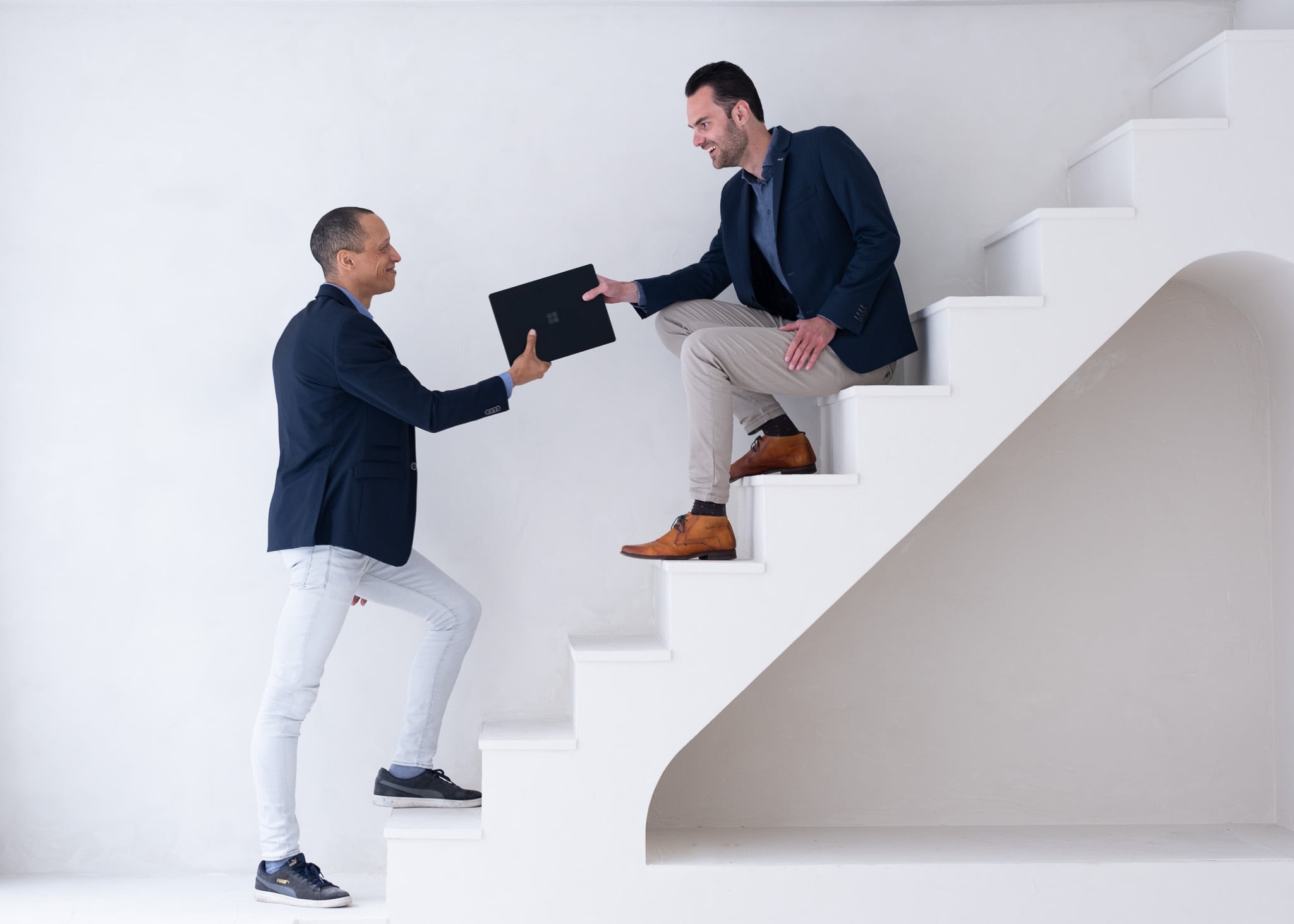 Twee zakelijke mannen op witte trap uitwisselend laptop in minimalistische studio setting