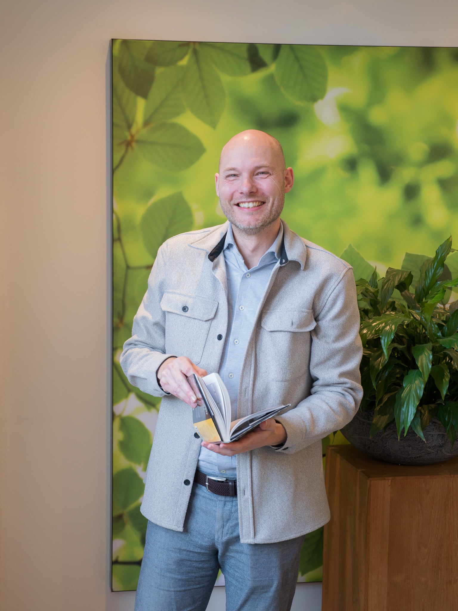 Glimlachende man in grijze jas bladert door boek voor groen bladerenbehang, staand bedrijfsportret met plant