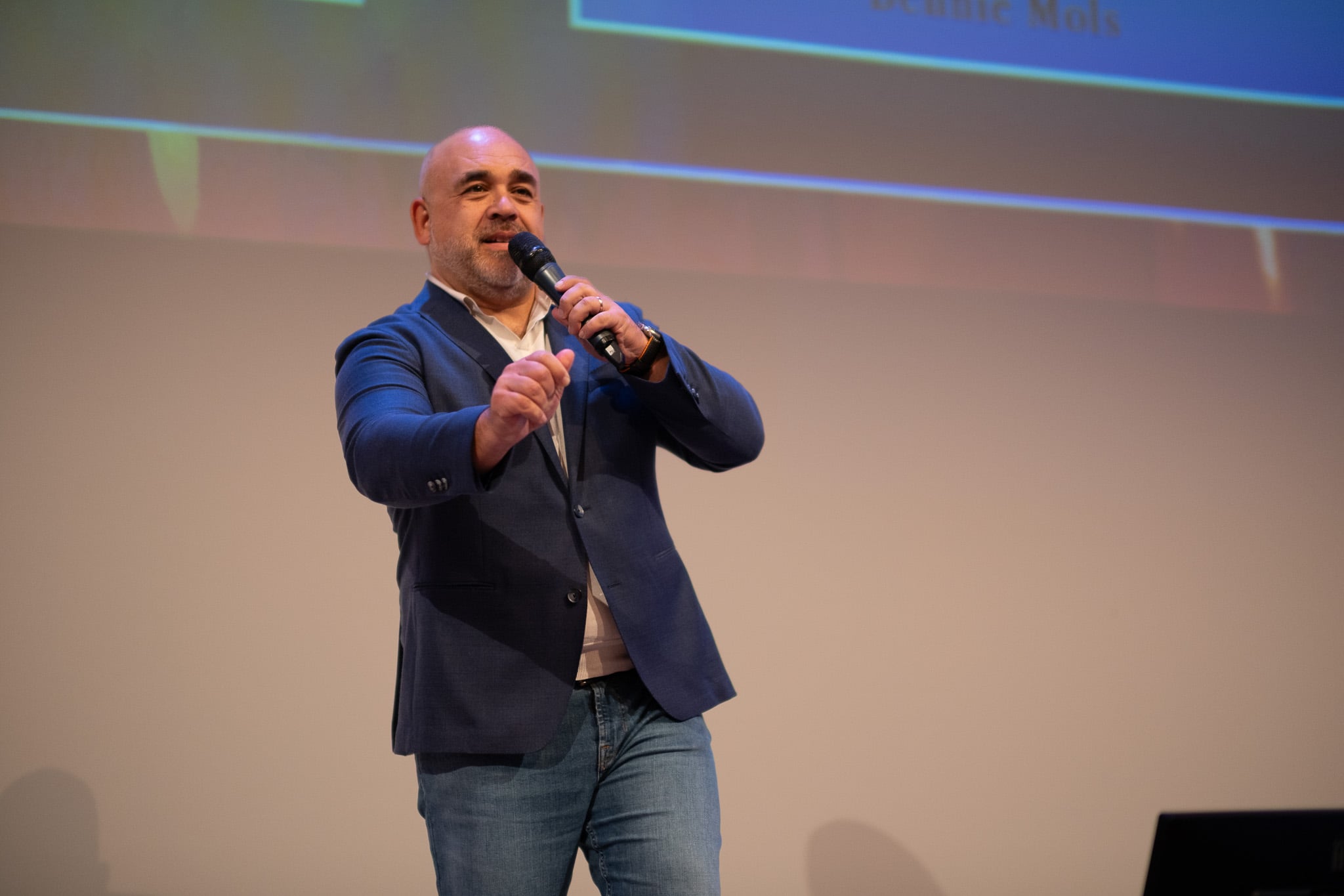 Kale spreker in donkerblauw pak presenteert enthousiast met microfoon op podium