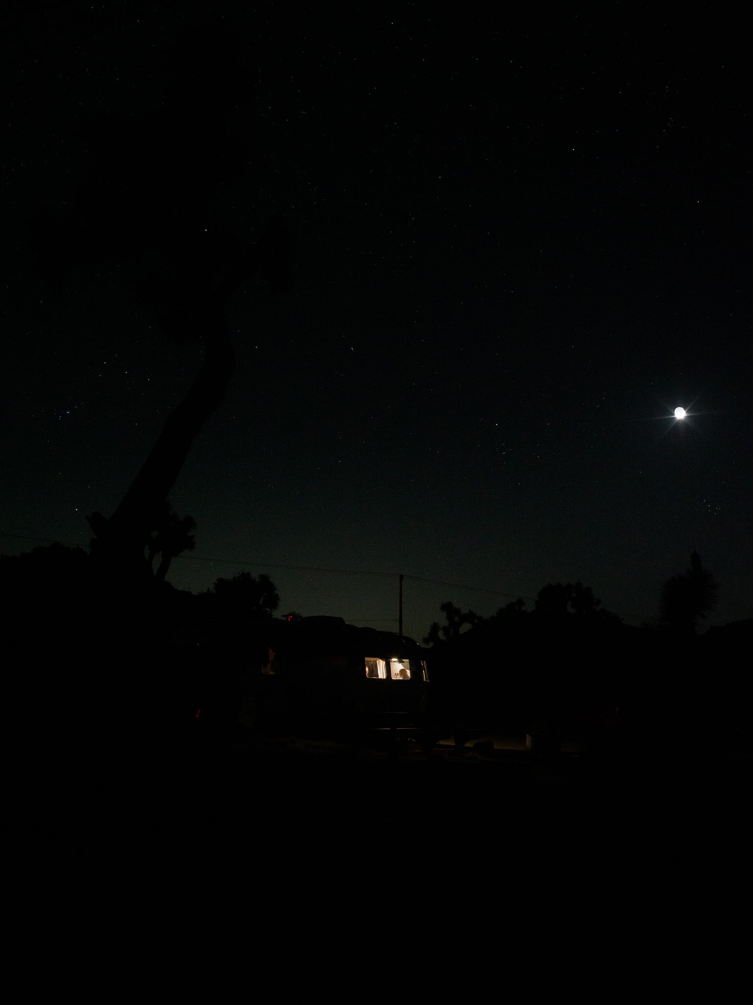 Silhouet van een verlichte caravan onder een sterrenhemel met heldere maan in de woestijnnacht