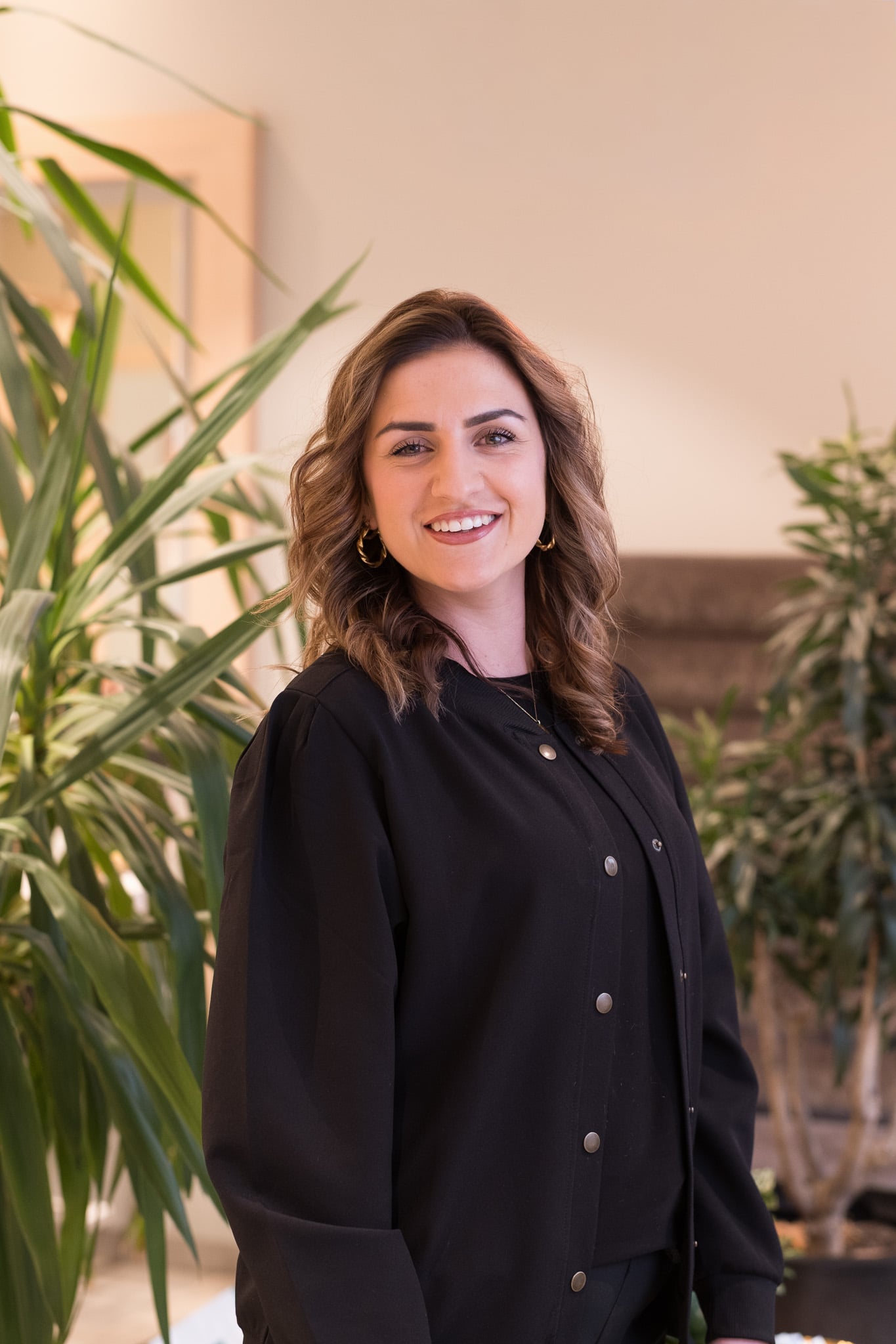 Brunette kapster met gouden oorringen in zwarte blazer tussen groene planten, warme natuurlijke belichting