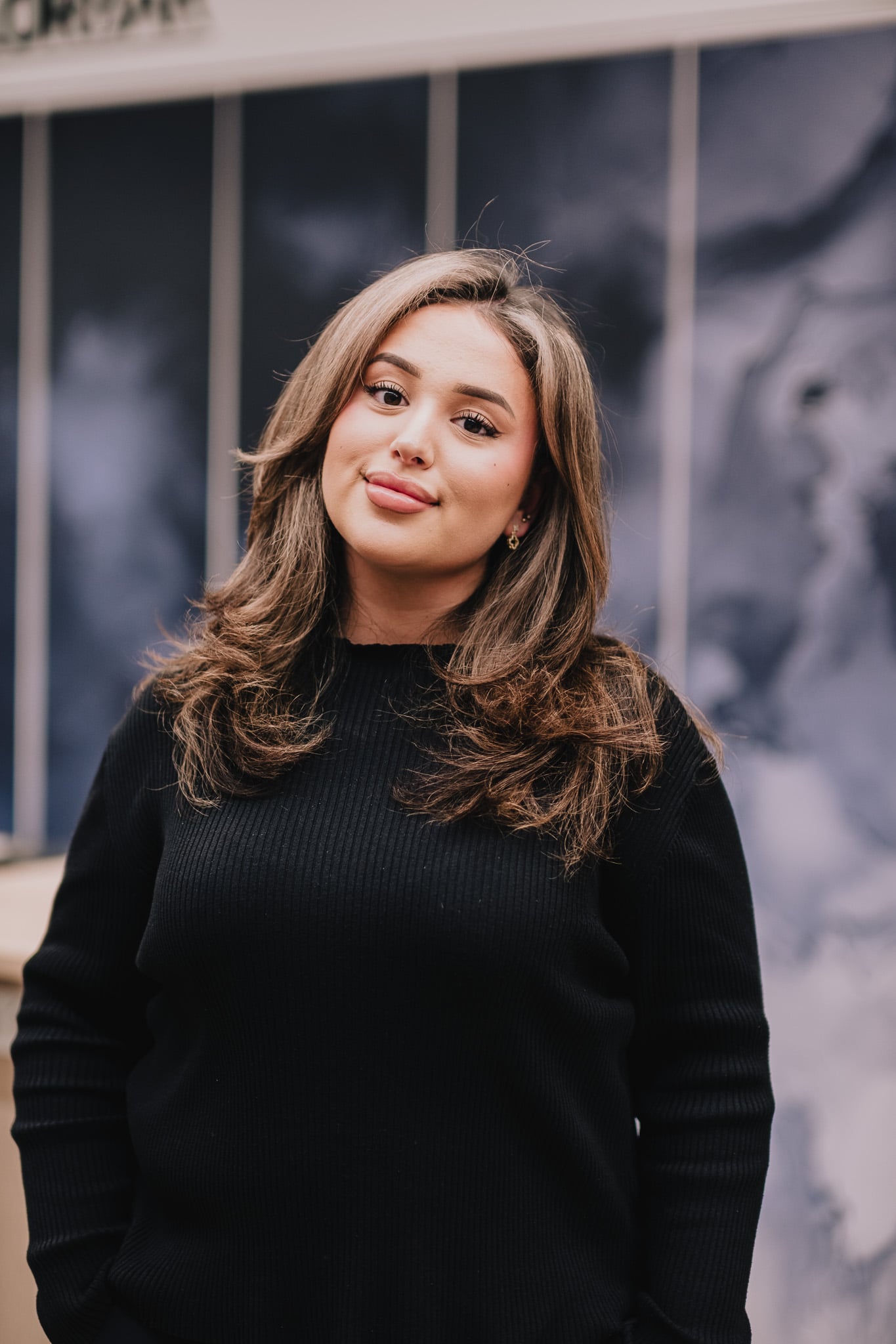 Jonge vrouw met lang bruin haar in zwarte coltrui glimlacht warm buiten kapsalon, natuurlijke portretfoto