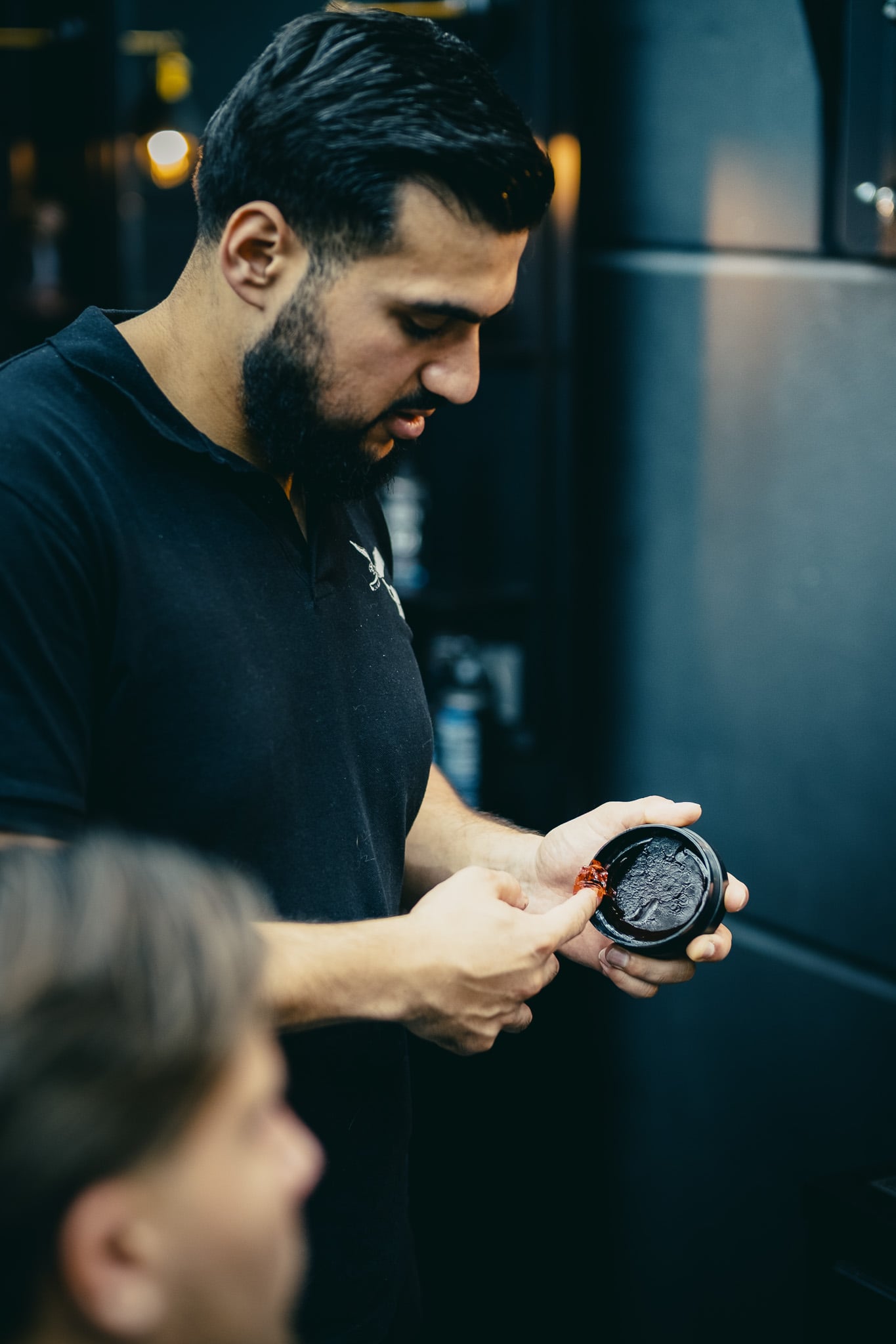 Barbier toont haarstyling product in donkere pot tijdens behandeling met warme studio verlichting