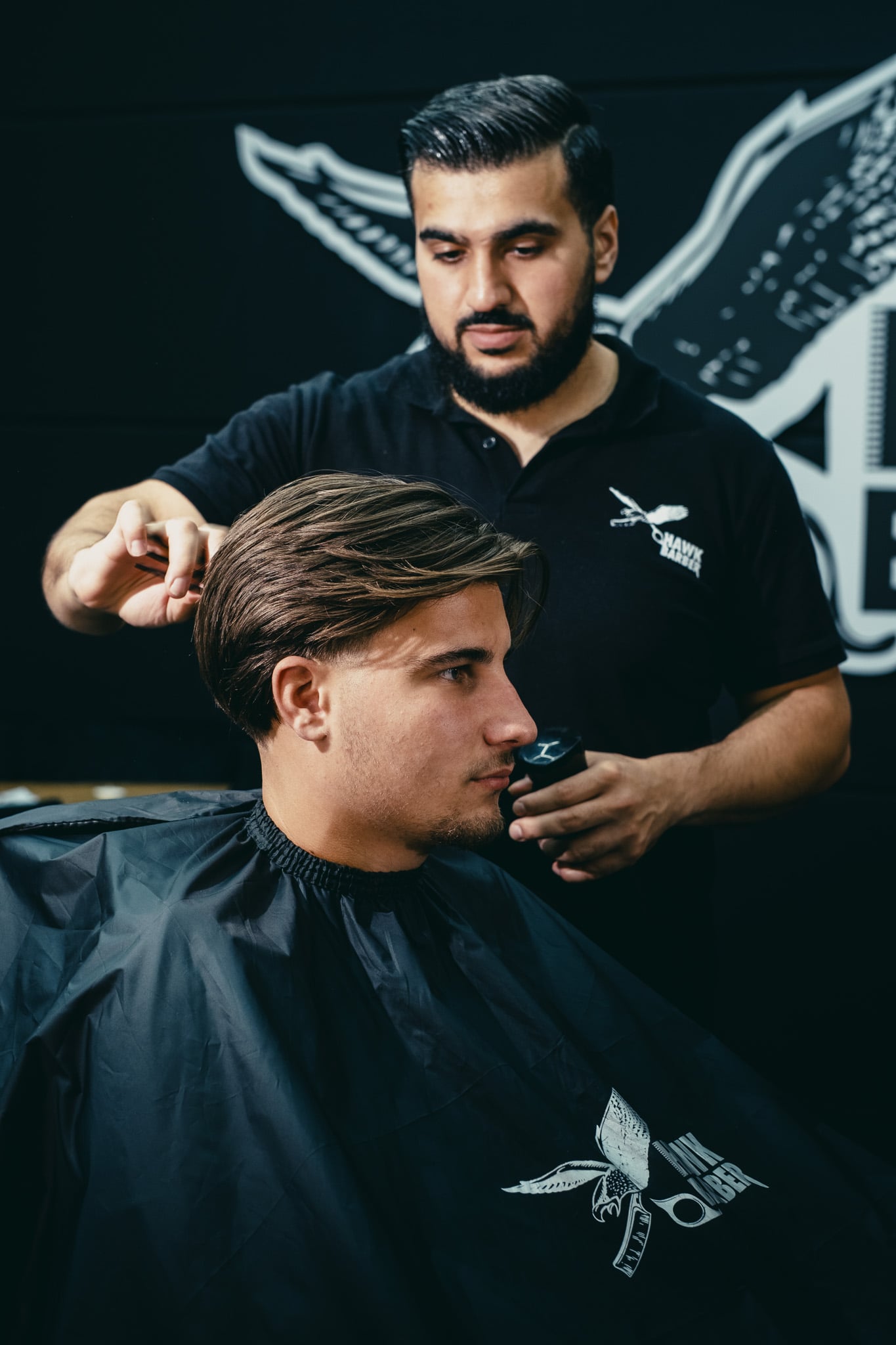 Ervaren kapper stijlt blond haar van klant met Hawk Barber cape in stijlvolle zaak