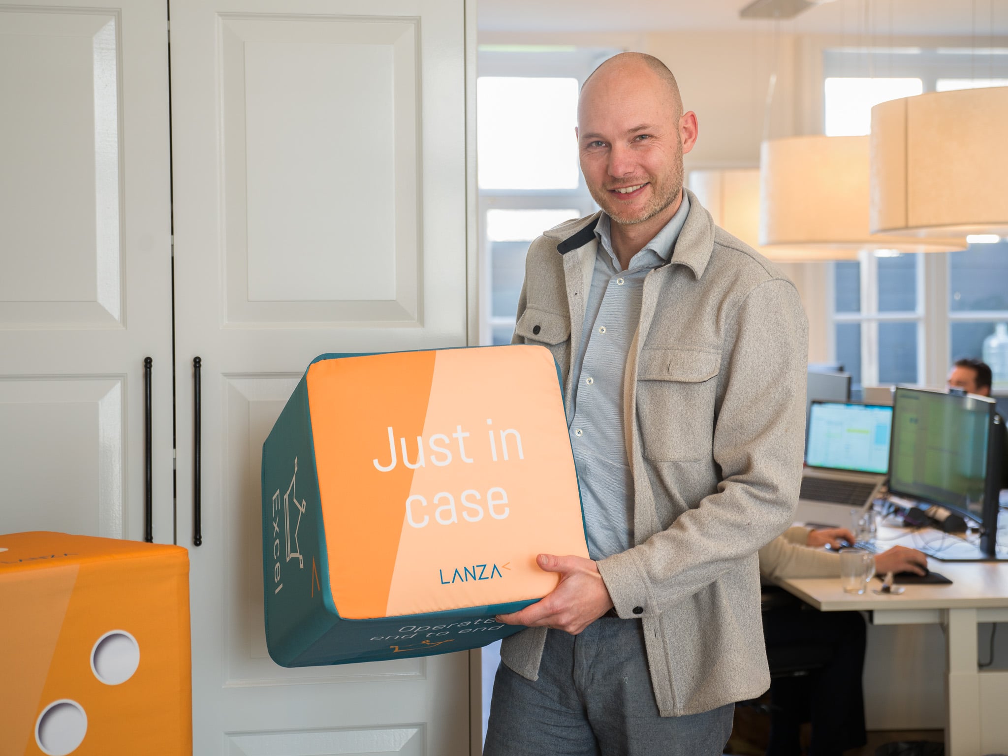 Man in wollen jasje houdt kussen met bedrijfslogo 'Just in case' vast in modern kantoor, zakelijk LinkedIn-portret