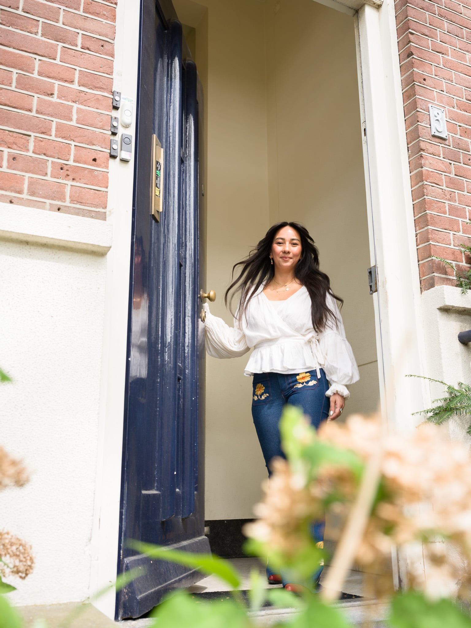 Vrouw met golvend donker haar en witte blouse loopt lachend naar buiten door blauwe voordeur, zakelijk portret in Amsterdamse stijl