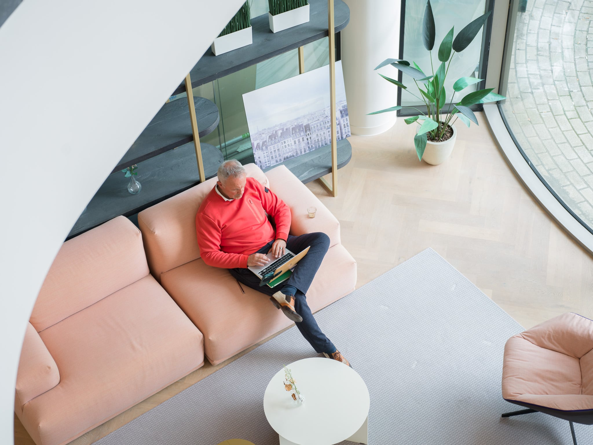 Vogelperspectief van man in rode trui werkend op laptop op roze bank in strak ingerichte kantoorlounge