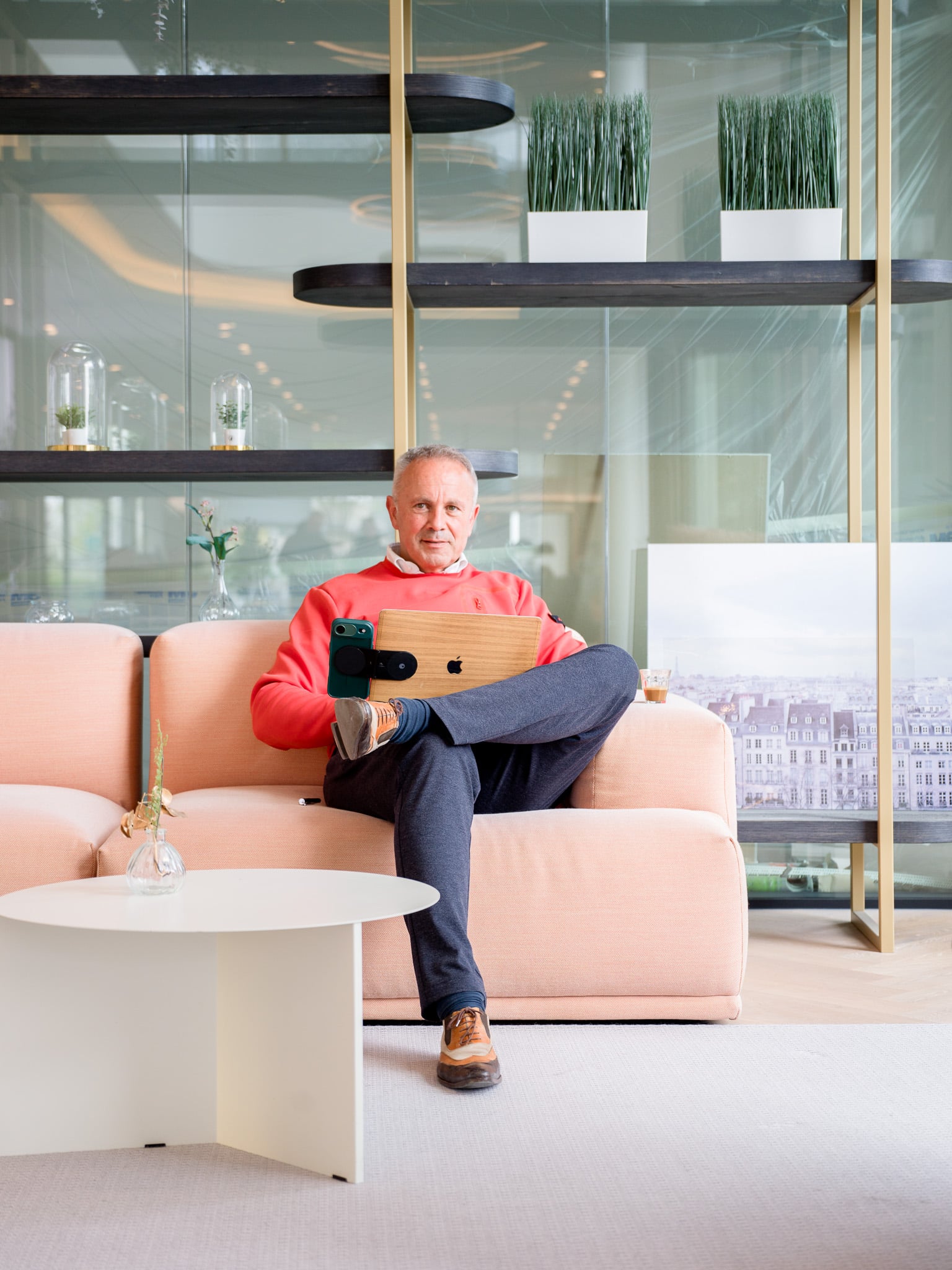 Man in koraalrode trui zit met gekruiste benen op roze bank en houdt laptop vast, zakelijk portret in luxe kantoorlobby