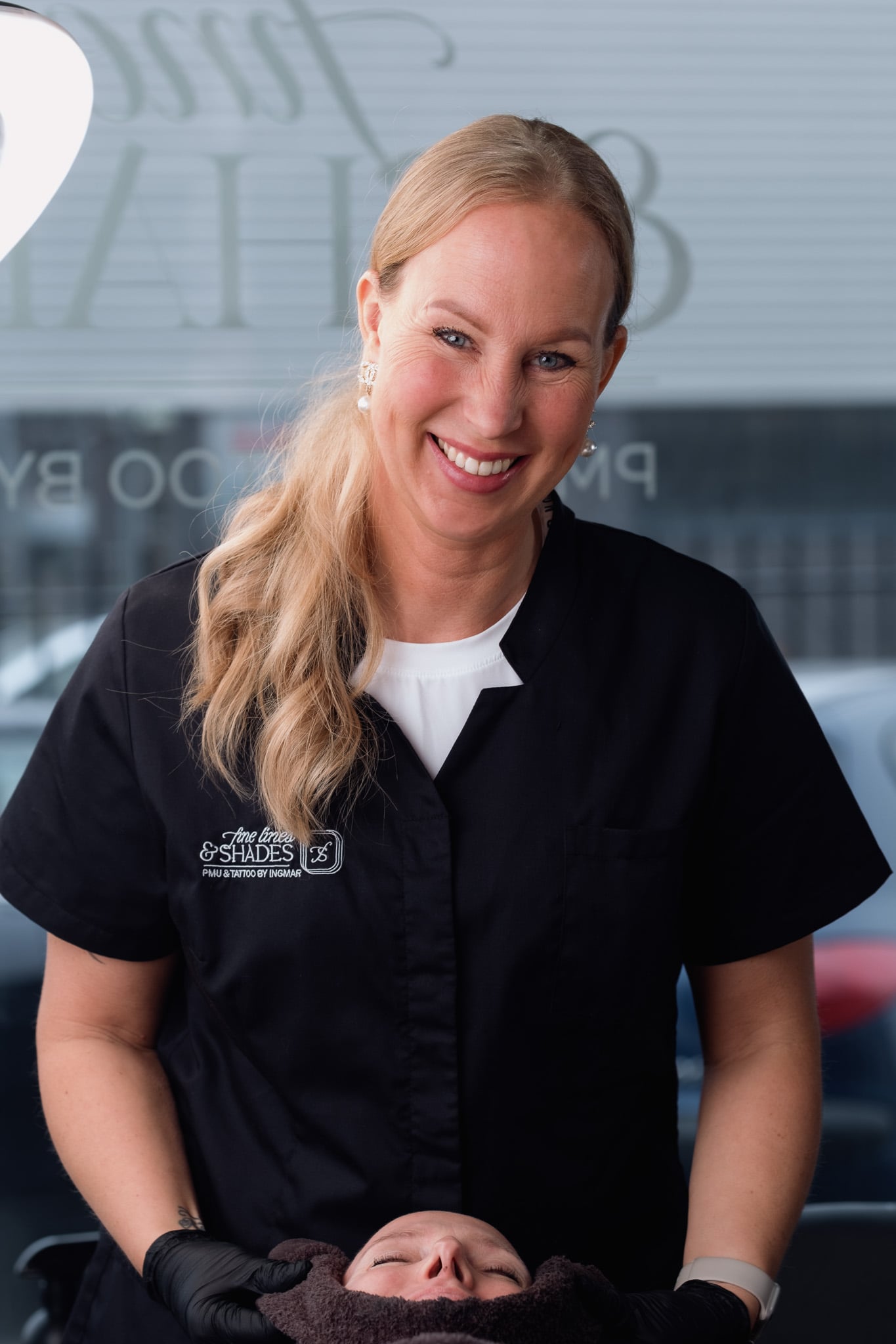 Lachende blonde schoonheidsspecialiste in zwart uniform poseert vriendelijk in moderne salon Fine Lines & Shades