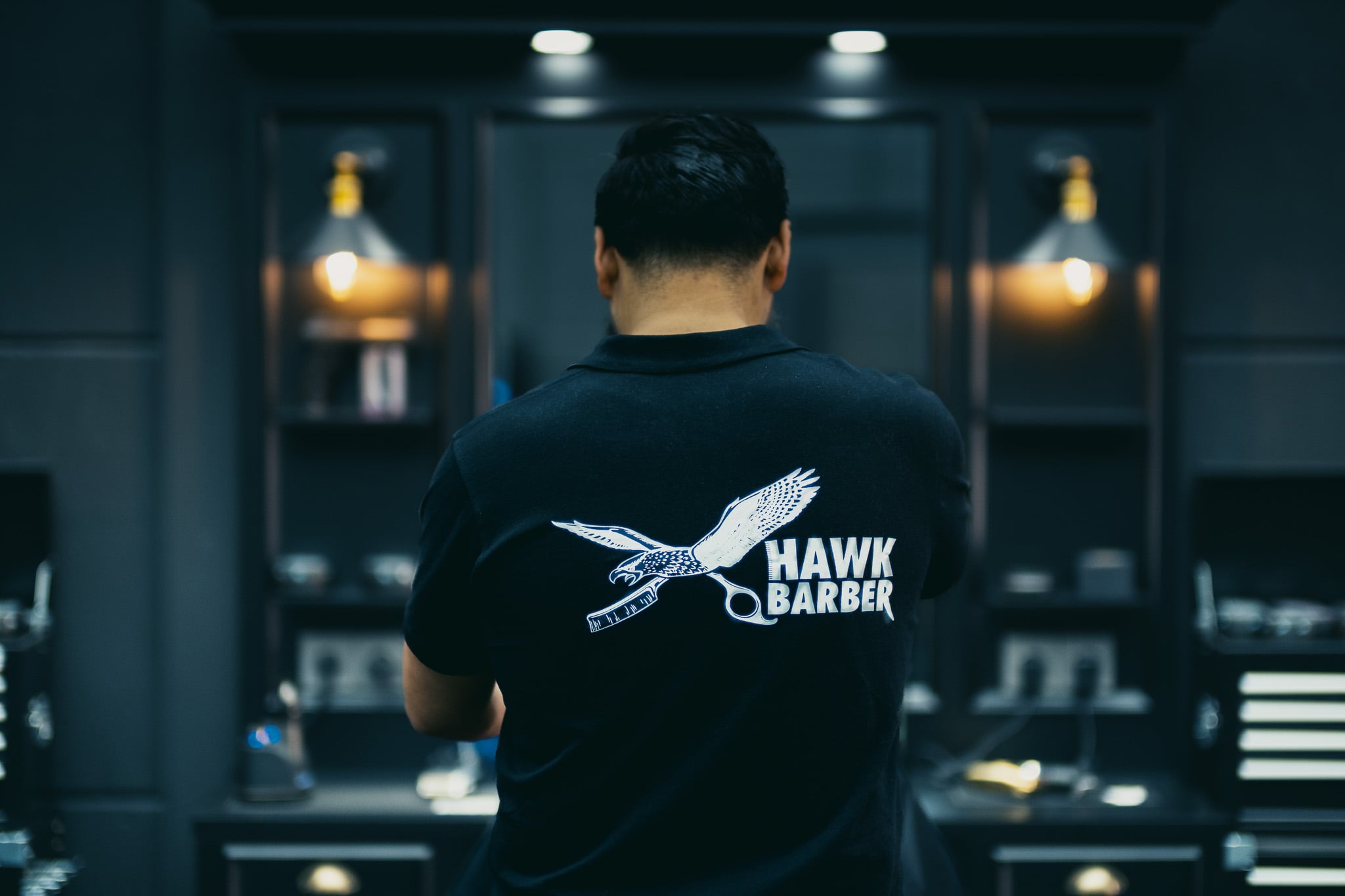 Barbier met Hawk Barber logo op rug toont professionele uitstraling in donkere barbershop interieur