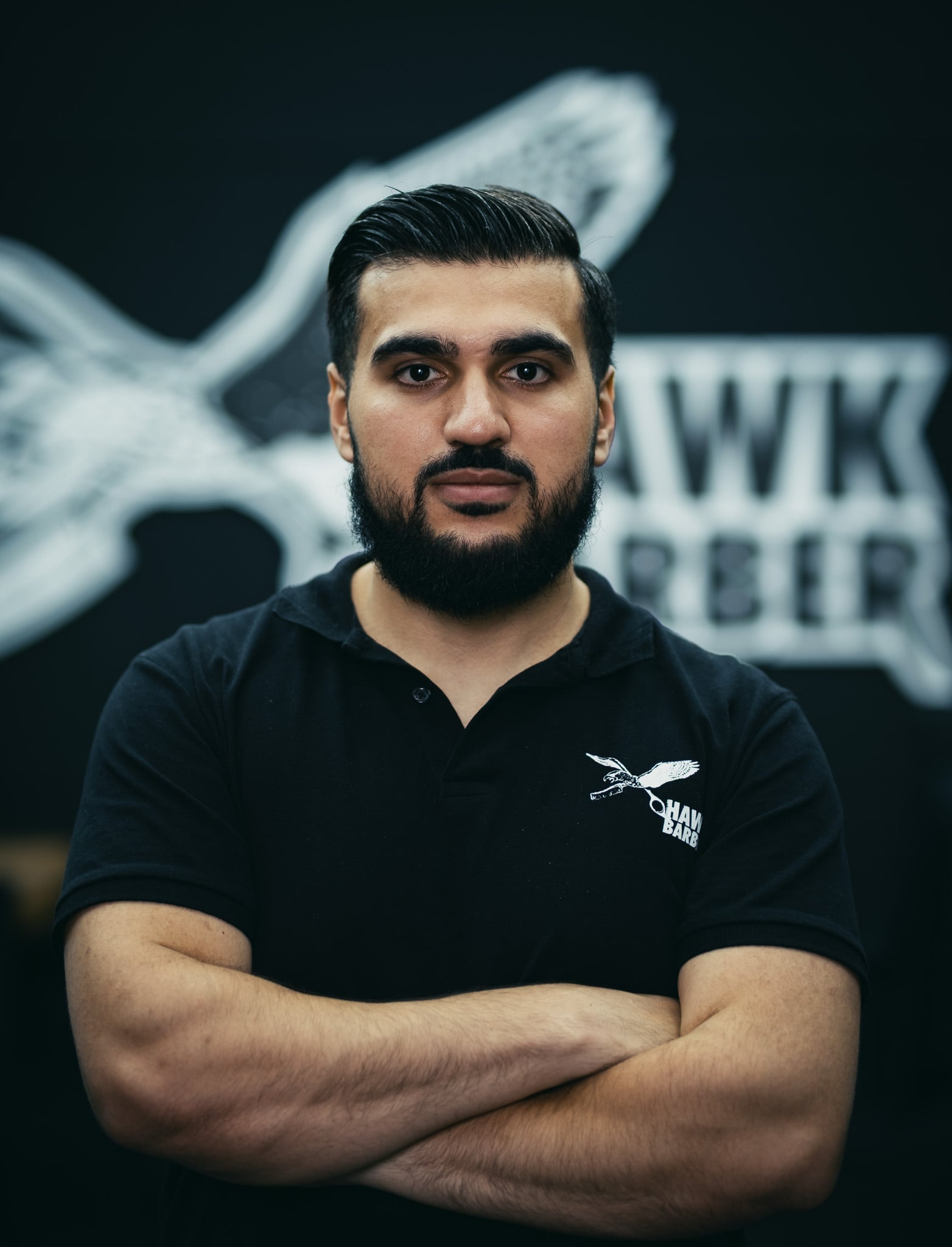 Zelfverzekerde barbier met gekruiste armen poseert voor Hawk Barber logo muur portret