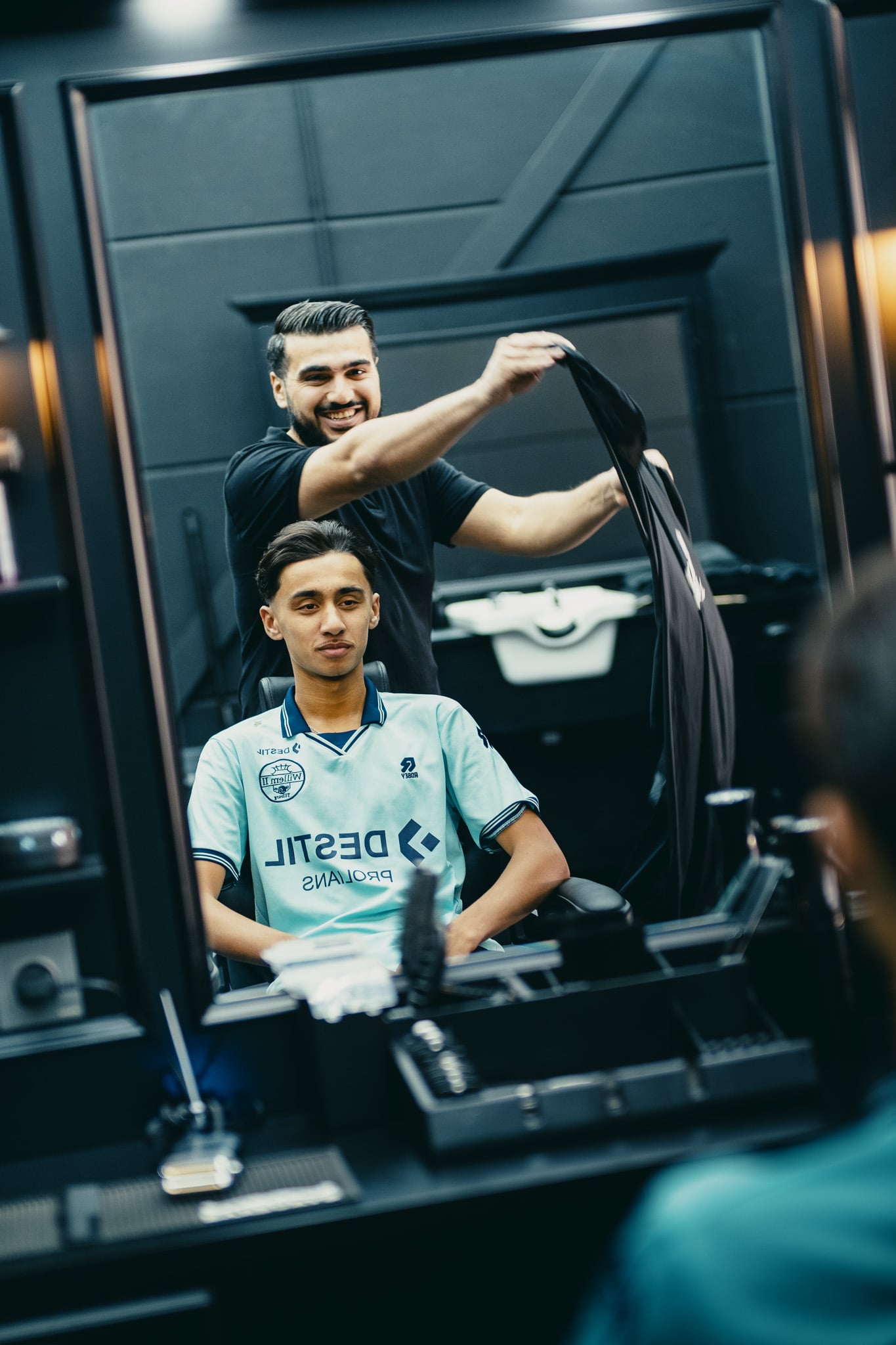 Vrolijke barbier in zwart uniform werkt aan klant in voetbalshirt bij moderne barbershop spiegel