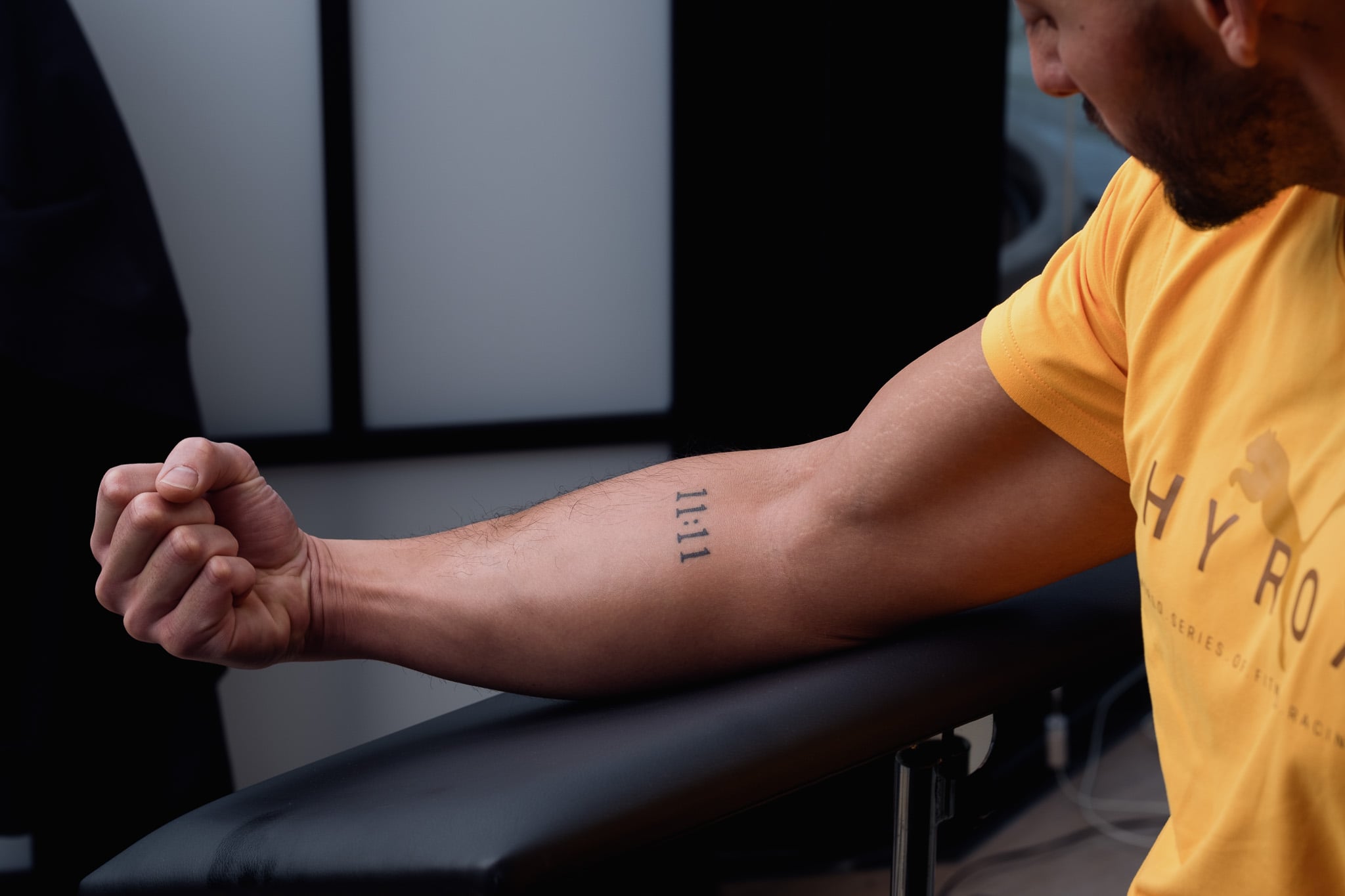Man in geel shirt toont verse tatoeage op onderarm tijdens PMU nabehandeling in schoonheidssalon