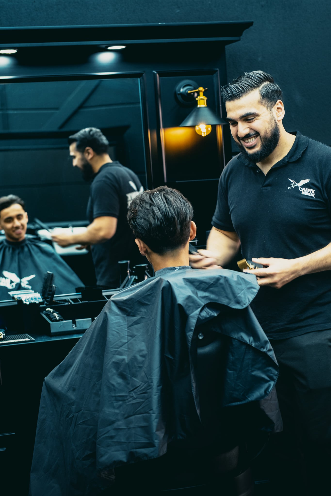 Lachende barbier in Hawk Barber poloshirt knipt haar van klant met warme lampensfeer op achtergrond