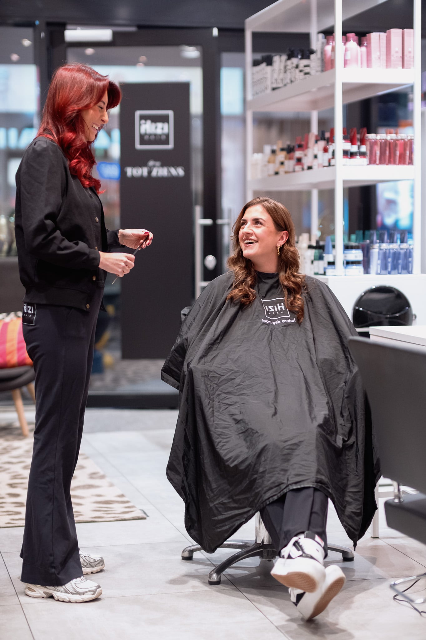 Roodharige kapster helpt lachende klant in kapsalonstoel, interactie bedrijfsfotografie Hizi Hair
